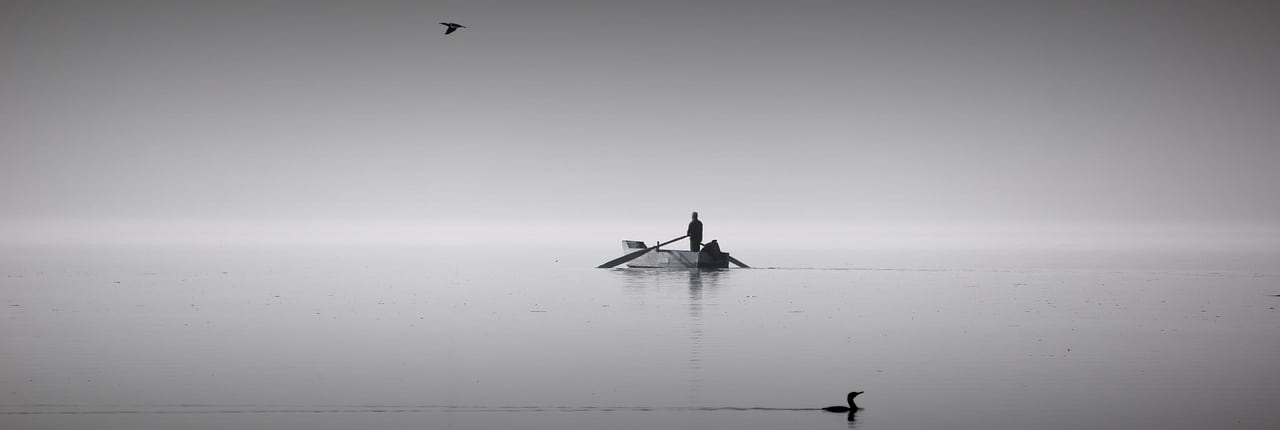 A reflective grayscale image capturing the serene minimalism of a lone individual paddling a small boat in calm waters, with birds silhouetted against a soft sky, suggesting peace and solitude.