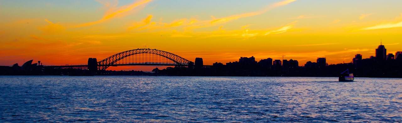 A scenic view of a sunset behind a silhouette of a large bridge, likely an iconic landmark, with the water in the foreground and the skyline of a city stretching across the horizon, under an orange and purple sky.